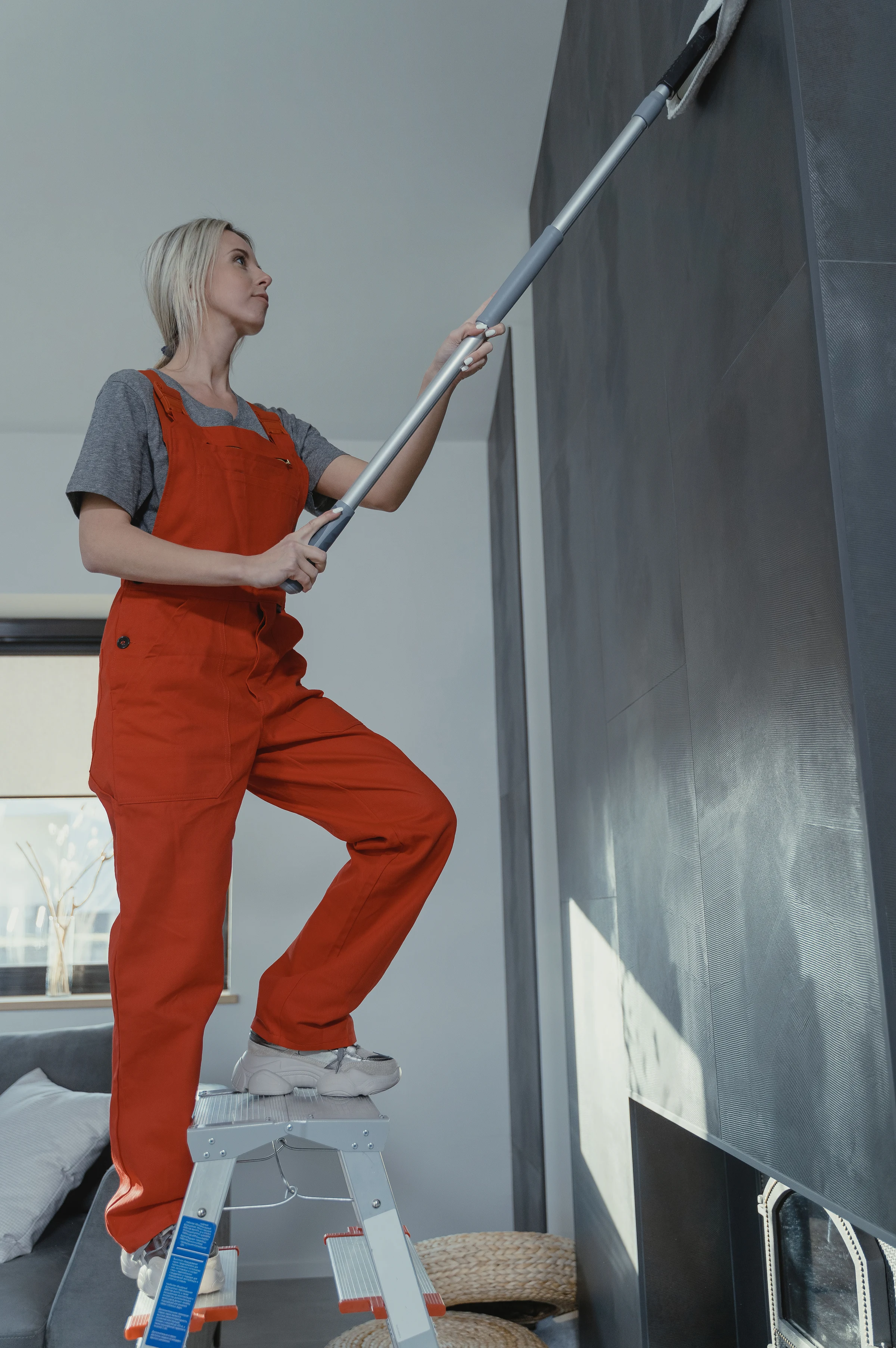 A cleaner on a step ladder dusting a high wall with an extension pole