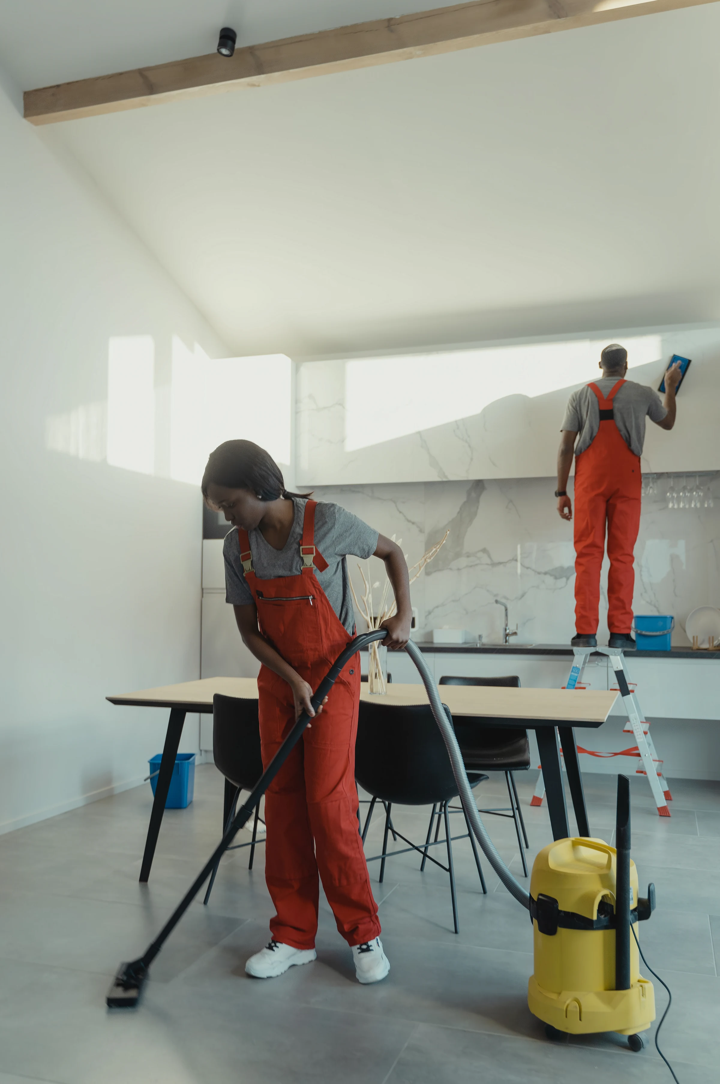 A cleaner vacuuming under a dining table while a colleague cleans the windows